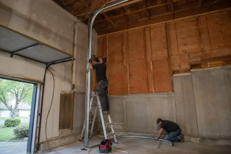 One person on a stepladder working beside a tall curved garage-door track while another kneels beside loose track sections in an unfinished garage. — High-Lift Garage Door Conversion, Guelph