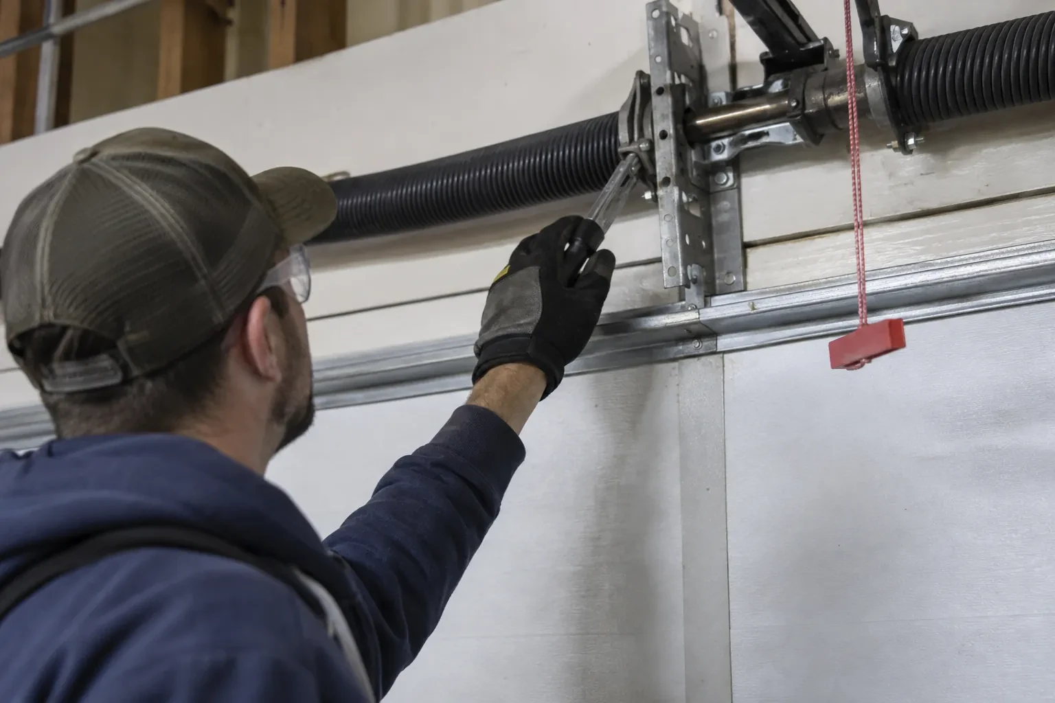 Technician in a cap reaching a gloved hand with a tool toward a coiled spring and bracket above a garage door. — Everton