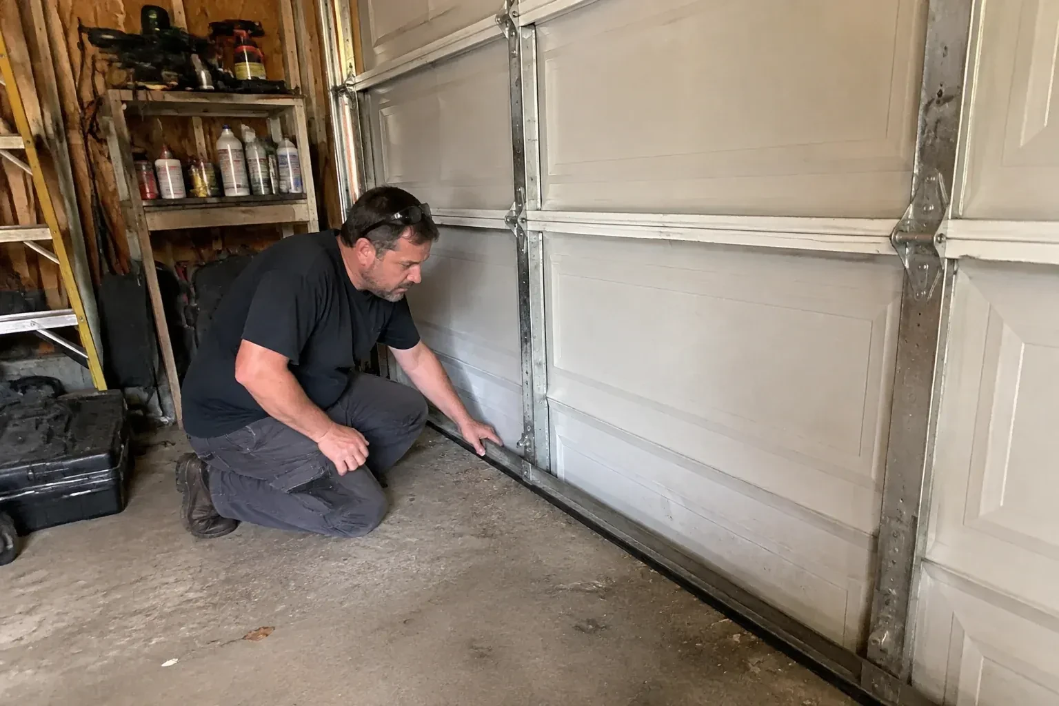 Person kneeling by a white paneled door in a garage, one hand on the black seal along its bottom edge. — Garage Door Roller Replacement, Guelph